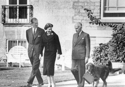 A black-and-white photo of The Queen and The Duke of Edinburgh walking outside Rideau Hall with Governor General Massey. A dog walks alongside them, carrying The Queen’s purse in its mouth. 