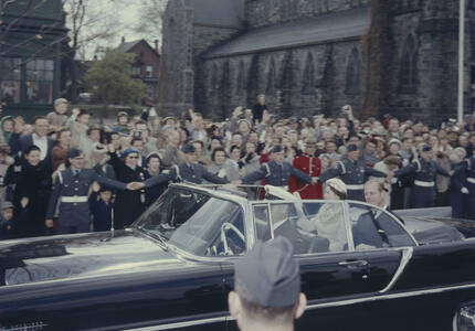 The Queen and the Duke of Edinburgh are being driven in a black convertible. A large crowd of people wave to them. A church stands in the background.