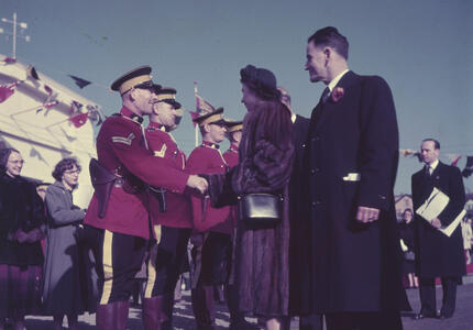 La princesse Elizabeth, qui porte un manteau de fourrure, serre la main d’un agent de la GRC, tandis que trois autres agents sont au garde-à-vous. Les agents portent des uniformes rouges. Un navire et des drapeaux nautiques sont à l’arrière-plan.