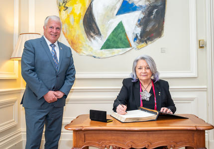 La gouverneure générale Mary Simon est assise à un bureau. Elle a un stylo à la main et signe un livre d'or. Un homme est debout à sa gauche. Ils sourient tous les deux.