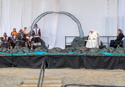 A large stage decorated with mountainous scenery. Performers are singing and playing instruments on the right side of the stage. Pope Francis and another man sit on the left side of the stage. 