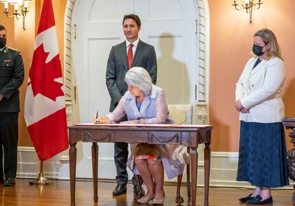 The Governor General is sitting at a desk signing a document. The Prime Minister is standing behind her.