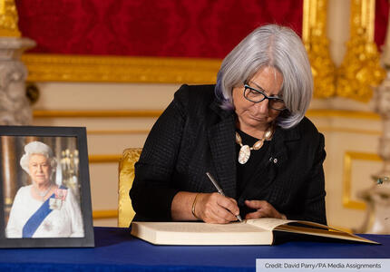 Governor General Simon is sitting at a table and writing in a book. There is a portrait of Her Majesty The Queen on the table. Text in the lower right corner of the image reads, “Credit: David Parry/PA Media Assignments.”