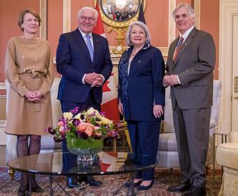 Governor General Simon Stands next to His Excellency Frank-Walter Steinmeier, President of the Federal Republic of Germany, Mr. Whit Fraser and Ms. Elke Büdenbende. They are smiling at the camera. Behind them is the flag of Canada and the flag of Germany.