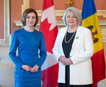 La gouverneure générale Mary Simon se tient à côté de la présidente de la Moldavie, Maia Sandu, à Rideau Hall. Les deux femmes sont souriantes. Un drapeau canadien et un drapeau moldave sont visibles derrière elles.