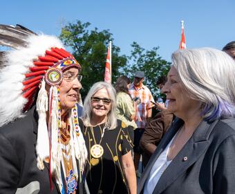 La gouverneure générale Mary Simon sourit et parle à un homme lors de la cérémonie de sélection du site du Monument national des pensionnats autochtones. Ils sont à l'extérieur par une journée ensoleillée. 