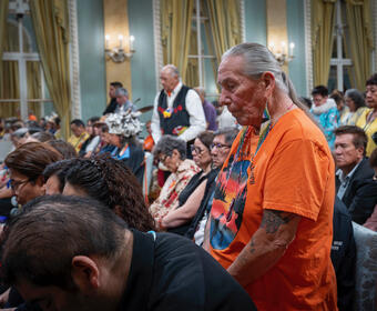 A silver haired man in an orange shirt stands among seated and standing peers.