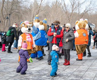 Governor General Simon and Mr. Fraser are smiling at a group of children. The Winterlude Ice Hogs and a group of people stand behind them.