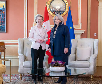 Governor General Simon Stands next to Her Excellency Ursula von der Leyen, President of the European Commission. They are smiling at the camera. Behind them is the flag of Canada and the flag of Europe.