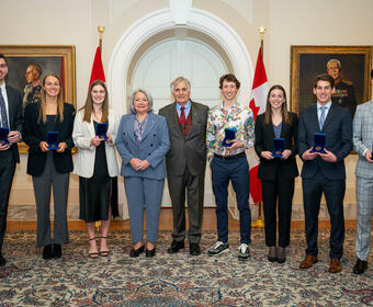 A group photo featuring Governor General Simon, Mr. Whit Fraser and seven recipients of the Governor General’s Academic All-Canadian Commendation. The recipients are each holding a blue box with a medallion inside of it. Behind them: two Canadian flags.