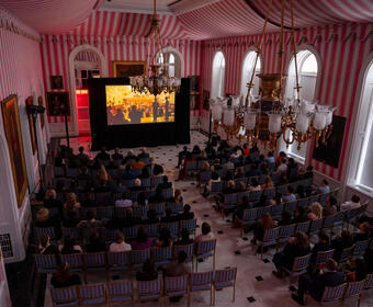 Une grande foule de personnes est assise dans la salle de la tente de Rideau Hall et regarde un film sur grand écran