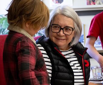  Governor General Mary Simon sits in a red chair, while holding a book and smiling at child who is standing beside her.