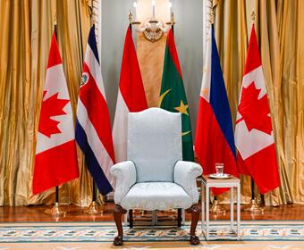 A regal chair flanked by flags from Canada, Costa Rica, Indonesia, Mauritania, and the Philippines. Elegant gold curtains create a formal, diplomatic ambiance.