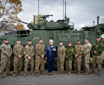 Governor General Mary Simon posing for a photo with a group of military personnel in camouflage uniforms and berets. A large army tank is behind them.