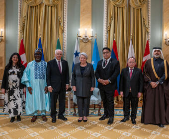 Governor General Simon standing with six&nbsp;new heads of mission, with the flags of each country behind them. Two Ceremonial Guards are in the back.