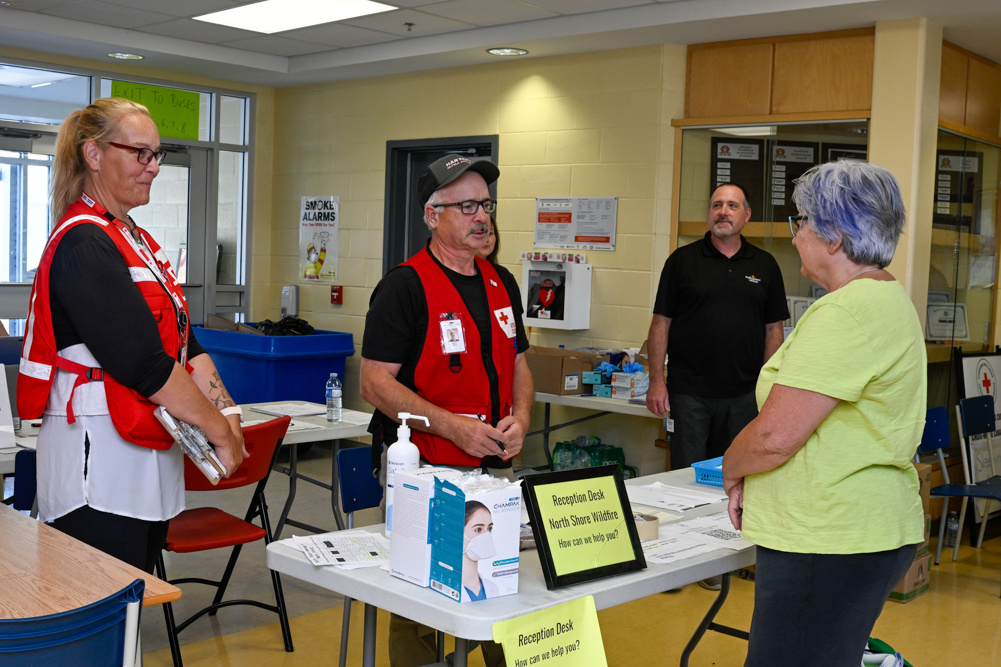 La gouverneure générale Mary Simon s'adresse à des bénévoles portant des gilets de la Croix-Rouge. Une affiche sur la table indique « Bureau d'accueil - Incendie de forêt de North Shore - Comment pouvons-nous vous aider ? ».